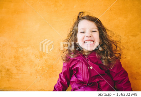 Young girl with a cheeky smile looking directly at the camera against a bright yellow wall background 136232949