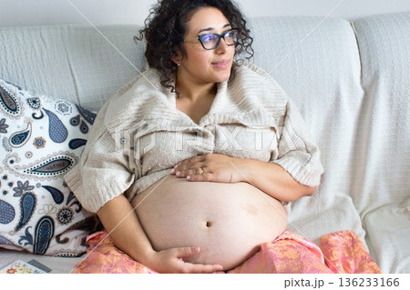 A young attractive white Caucasian woman sitting on a sofa at home, rubbing her pregnant belly, looking contemplative and thoughtful A young attractive white Caucasian woman sitting on a sofa at home, rubbing her pregnant belly, looking contemplative and thoughtful 136233166