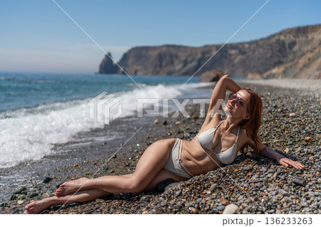 Beautiful woman bikini sunbathing pebble beach cliff ocean waves vacation relaxation 136233263