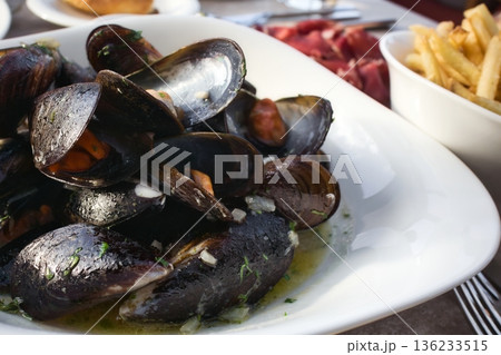 Close-up of a big white ceramic plate of black shell mussels in a white wine garlic sauce with french fries in the background 136233515