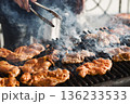 Close-up shot of a white Caucasian's mas holding metal tongs behind a charcoal barbeque with sizzling pieces of open chicken thighs 136233533