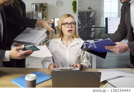 Overwhelmed female employee swamped with tasks as colleagues crowd desk with files 136234443