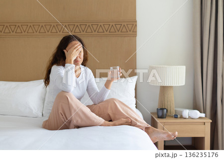 Woman at home sitting on bed holding glass of water, feeling unwell Woman at home sitting on bed holding glass of water, feeling unwell 136235176