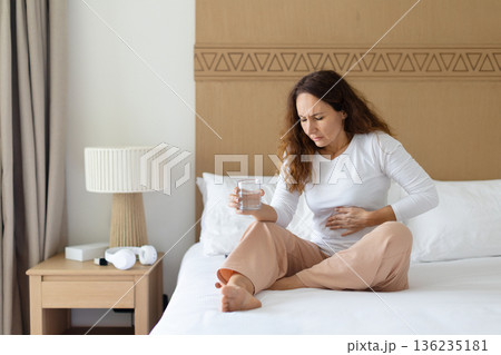 Woman sitting on bed at home holding stomach and glass of water Woman sitting on bed at home holding stomach and glass of water 136235181
