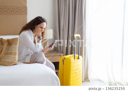 Woman sitting on bed at home, looking worried at smartphone beside suitcase 136235250