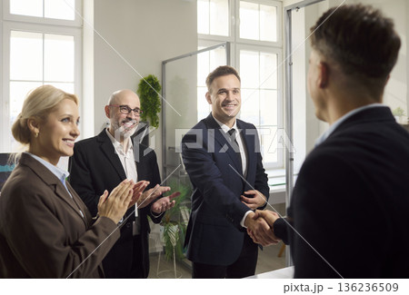 Businessmen handshake in office meeting as colleagues applaud new partnership deal Businessmen handshake in office meeting as colleagues applaud new partnership deal 136236509