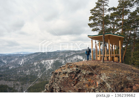 Man with trekking poles stands by a wooden gazebo on a mountain cliff, overlooking a winter forest. Hiking concept 136239662