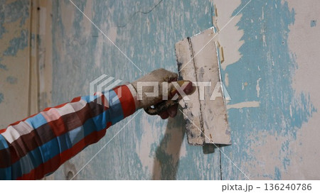 scraping off the remains of wallpaper from peeling walls with a wide trowel, a close-up of a hand in a plaid shirt and a dirty glove with a spatula near the wall of a room, picking off scraps scraping off the remains of wallpaper from peeling walls with a wide trowel, a close-up of a hand in a plaid shirt and a dirty glove with a spatula near the wall of a room, picking off scraps 136240786