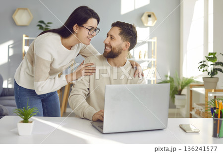 Happy loving woman looking at her husband while he is sitting at his working desk with his laptop Happy loving woman looking at her husband while he is sitting at his working desk with his laptop 136241577