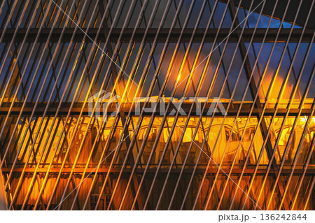 Modern architecture facade with diagonal metal framework, reflective glass panels illuminated by warm interior light, geometric pattern. Abstract architecture background, urban design, copy space 136242844