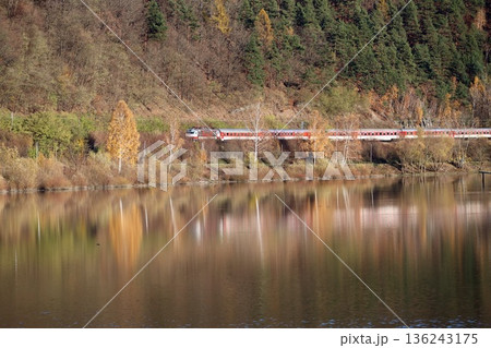 a red passenger train traveling along a calm lake. a red passenger train traveling along a calm lake. 136243175