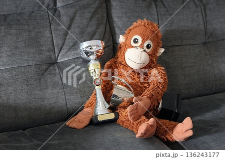 A stuffed monkey is sitting on a couch holding two cups and two trophies 136243177