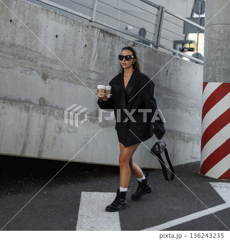 Woman in oversized black blazer and sunglasses holding takeaway coffee cups, walking in city street with confidence and style. Beautiful urban lady with coffee near concrete wall 136243235