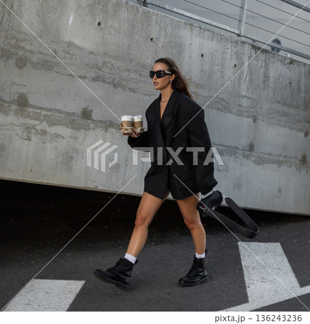 Fashionable young woman in oversized black blazer and sunglasses holding takeaway coffee cups, walking outdoors with confidence and modern street style. Beautiful urban lady with coffee near concrete 136243236