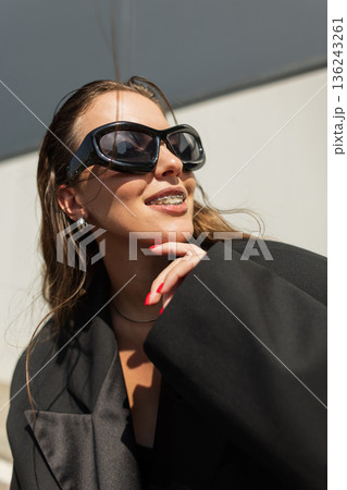 Fashionable young woman smiling in sunglasses and black jacket. Stylish model in oversized blazer and shades enjoying sunny day, posing with a playful gesture, perfect for lifestyle, streetwear Fashionable young woman smiling in sunglasses and black jacket. Stylish model in oversized blazer and shades enjoying sunny day, posing with a playful gesture, perfect for lifestyle, streetwear 136243261