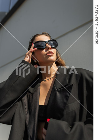 Young female model in trendy outfit with oversized jacket and accessories, posing outdoors in sunlight. Cool beautiful girl in black clothes puts on sunglasses in the city 136243271