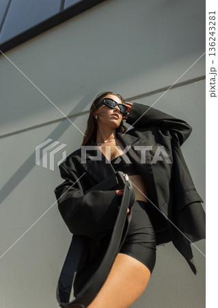 Confident oung woman in fashionable black outfit and sunglasses looking up under bright sunlight. Contemporary street style portrait. Confident oung woman in fashionable black outfit and sunglasses looking up under bright sunlight. Contemporary street style portrait. 136243281