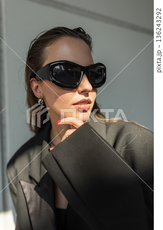 Young woman in oversized black blazer and dark sunglasses posing outdoors, modern street style fashion portrait with confident and trendy look. 136243292