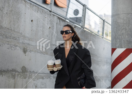 Fashionable woman in oversized black blazer and sunglasses carrying takeaway coffee cups, walking confidently in urban street setting. 136243347