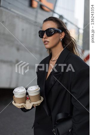 Confident woman in black blazer dress with coffee-to-go, stylish sunglasses and sleek hairstyle, urban lifestyle. Beautiful urban lady with coffee near concrete wall Confident woman in black blazer dress with coffee-to-go, stylish sunglasses and sleek hairstyle, urban lifestyle. Beautiful urban lady with coffee near concrete wall 136243354