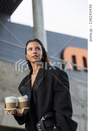 Trendy woman wearing oversized blazer carrying takeaway drinks, showcasing minimalist fashion and contemporary urban lifestyle. Trendy woman wearing oversized blazer carrying takeaway drinks, showcasing minimalist fashion and contemporary urban lifestyle. 136243430
