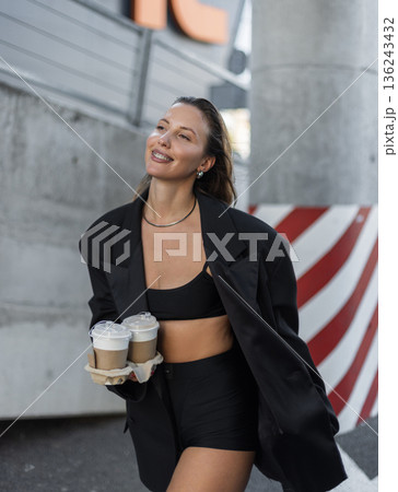 Smiling young woman in black blazer and crop top holding takeaway coffee cups outdoors, showing modern street fashion, confidence, and active urban lifestyle. 136243432