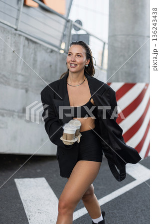 Smiling woman in trendy black outfit holding takeaway coffee cups while walking outdoors, showcasing modern street fashion and positive vibes. 136243438