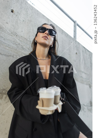 Stylish woman in oversized black blazer and sunglasses carrying coffee cups, stands near concrete wall in the city. Pretty business urban lady 136243452