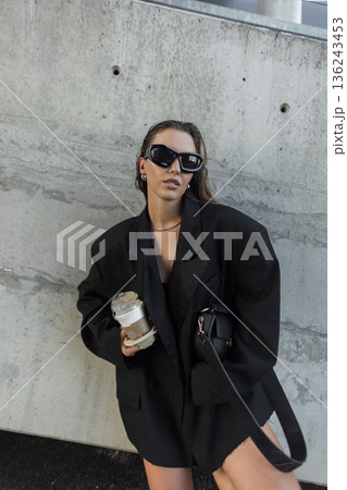 Trendy young woman holding two takeaway coffee cups, dressed in chic black blazer and modern sunglasses stands near concrete wall. Trendy young woman holding two takeaway coffee cups, dressed in chic black blazer and modern sunglasses stands near concrete wall. 136243453
