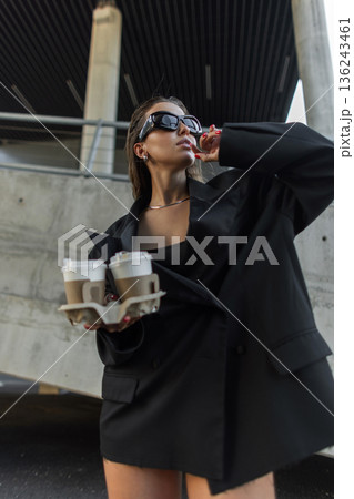 Chic woman enjoying urban lifestyle, wearing sunglasses and oversized blazer while holding coffee cups. Chic woman enjoying urban lifestyle, wearing sunglasses and oversized blazer while holding coffee cups. 136243461