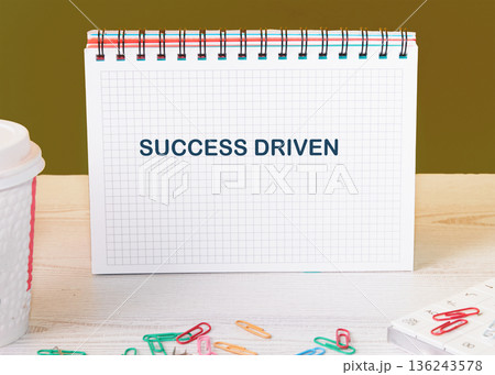 A spiral notebook on a light wooden desk shows SUCCESS DRIVEN. A white cup and colorful paper clips are near it, against an olive green wall A spiral notebook on a light wooden desk shows SUCCESS DRIVEN. A white cup and colorful paper clips are near it, against an olive green wall 136243578