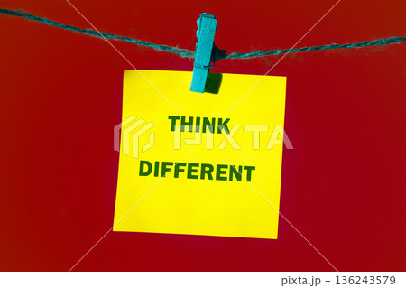 A yellow sticky note displaying the motivational phrase THINK DIFFERENT in green letters hangs from a blue clothespin on a string over a red background A yellow sticky note displaying the motivational phrase THINK DIFFERENT in green letters hangs from a blue clothespin on a string over a red background 136243579
