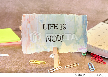 A piece of torn paper displays the motivational message LIFE IS NOW, held by a wooden clothespeg A piece of torn paper displays the motivational message LIFE IS NOW, held by a wooden clothespeg 136243580