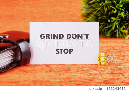 A white card reading GRIND DONT STOP stands on a textured orange surface, held by a yellow clip. A small dark book and green plant are nearby A white card reading GRIND DONT STOP stands on a textured orange surface, held by a yellow clip. A small dark book and green plant are nearby 136243611