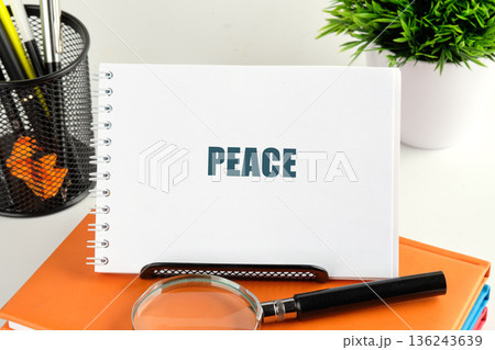A white spiral notebook showing the word PEACE is on orange books. A magnifying glass, pen holder, and green plant complete the calm desk scene 136243639
