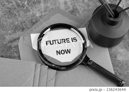 A magnifying glass rests on paper showing the words FUTURE IS NOW. Brown envelopes and a dark diffuser stand on a textured grey desk surface 136243649