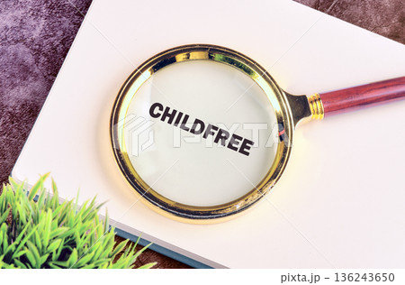 A gold-rimmed magnifying glass with a wooden handle rests on a white notebook, clearly enlarging the bold word CHILDFREE. Green foliage is visible on the side 136243650