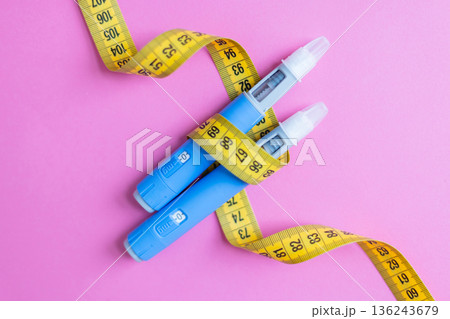 Two blue insulin pens wrapped with a yellow measuring tape on a pink background, symbolizing diabetes management, weight loss, medical treatment, and healthcare concepts. drugs injection ozempic 136243679