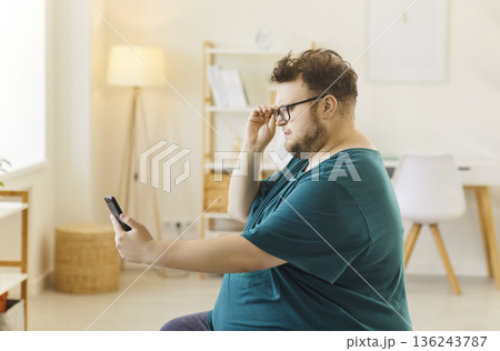 Plus-size man adjusting glasses while using smartphone at home on the sofa 136243787