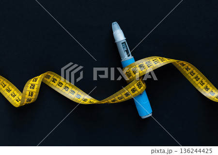 Blue insulin pen wrapped with a yellow measuring tape on black background. Symbol of diabetes, weight loss, obesity, or health management. Medical and lifestyle concept 136244425