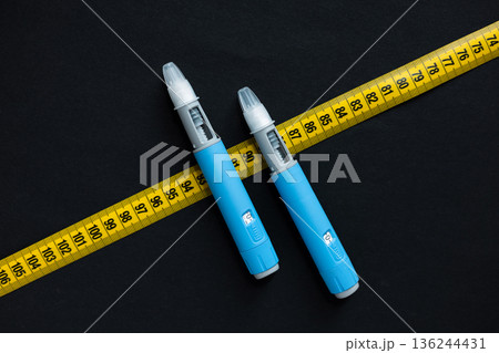 Blue insulin pen entangled in a measuring tape on black surface. Metaphor for weight-related illnesses, diabetic care, and modern medical treatment Blue insulin pen entangled in a measuring tape on black surface. Metaphor for weight-related illnesses, diabetic care, and modern medical treatment 136244431
