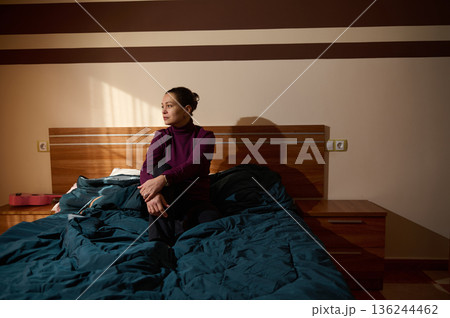 Woman Sitting On Bed In Dim Bedroom With Purple Turtleneck And Blue Blanket 136244462