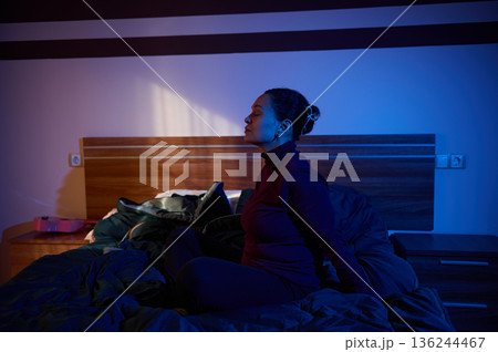 Relaxed Woman Sitting on Bed in Dim Blue Lighting For Quiet Night At Home 136244467