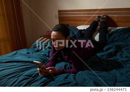 Woman Lying On Bed Using Smartphone In Cozy Dimly Lit Bedroom At Night 136244471