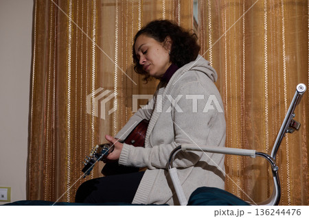 Woman Playing Acoustic Guitar While Sitting On A Bed In Cozy Apartment Setting 136244476