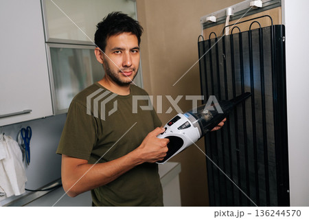 Portrait of young man cleaning refrigerator back coils with handheld vacuum, removing dust buildup for improved cooling efficiency and lower energy consumption in modern kitchen setting Portrait of young man cleaning refrigerator back coils with handheld vacuum, removing dust buildup for improved cooling efficiency and lower energy consumption in modern kitchen setting 136244570