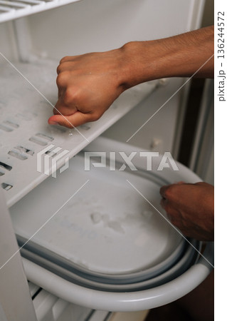 Vertical cropped shot of unrecognizable man squeezing melting ice cubes from refrigerator ice maker tray, hands wet from the task of defrosting freezer, water dripping frozen cubes removed. Vertical cropped shot of unrecognizable man squeezing melting ice cubes from refrigerator ice maker tray, hands wet from the task of defrosting freezer, water dripping frozen cubes removed. 136244572