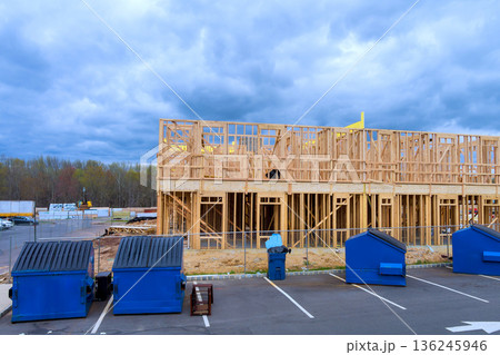 Workers build new apartment structure with wooden beams framing while dumpsters stand in parking area nearby. 136245946