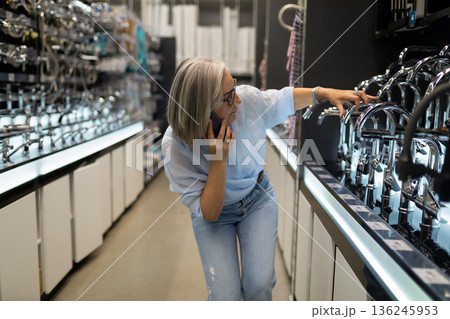 Woman examines kitchen faucets while on phone in a home improvement store during daylight Woman examines kitchen faucets while on phone in a home improvement store during daylight 136245953