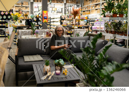 Woman sitting on a sofa in a store filled with plants and home decor enjoying her phone in a busy 136246066